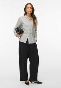 Femme portant une chemise blanche ample à rayures avec boutons, un pantalon large noir, des talons noirs à bout pointu, tenant un petit sac noir sur fond blanc.