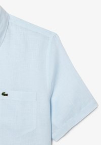 Camicia corta azzurra in lino, con taschino sul petto e un piccolo logo di coccodrillo verde. Tessuto strutturato con un design casual.