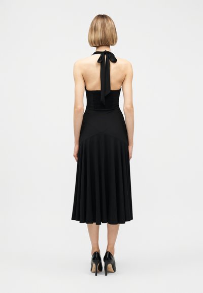 Femme aux cheveux blonds courts portant une robe noire à col licou avec un nœud papillon au cou et des talons hauts noirs, debout de dos.