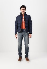 Jachetă puffer marină cu guler în picioare, fermoar frontal și textură matlasată. Poartă o bluză de culoare portocalie vibrantă, asortată cu blugi ușor albaștri și adidași maro.