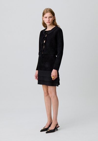 Vestido de ante negro con un corsé ajustado, falda de textura en capas y escote redondo con borde trenzado. Destacado con un lazo de cinta en la parte delantera.