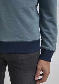 Blågrøn trøje med ribbede marineblå manchetter og kant, blød tekstur og en let tætsiddende pasform, parret med mørkegrå jeans.
