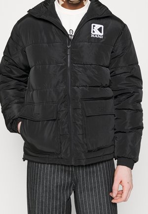 Personne portant une doudoune noire avec poches avant et logo Kani, associée à un pantalon noir à fines rayures et un t-shirt blanc à motif.