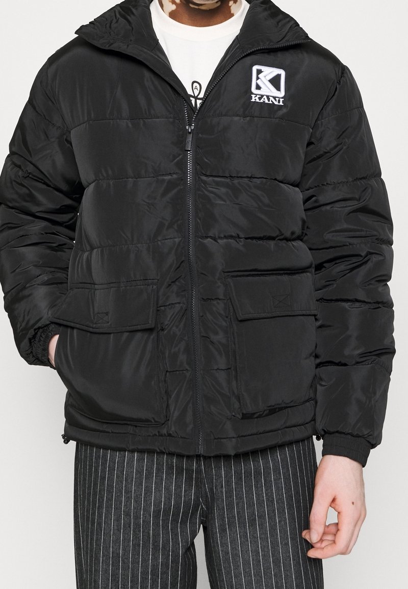 Personne portant une doudoune noire avec poches avant et logo Kani, associée à un pantalon noir à fines rayures et un t-shirt blanc à motif.
