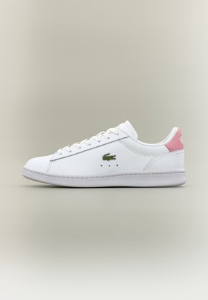 Baskets basses blanches avec lacets blancs, logo crocodile vert sur le côté et languette de talon en daim rose sur fond neutre.