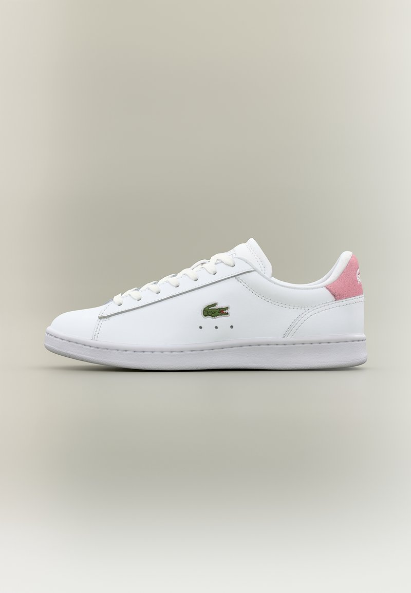 Baskets basses blanches avec lacets blancs, logo crocodile vert sur le côté et languette de talon en daim rose sur fond neutre.