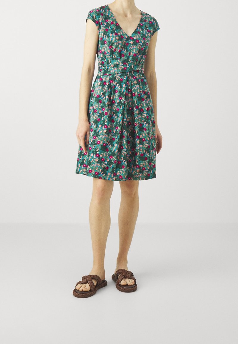 Sukienka w kwiaty z dekoltem w szpic, krótkimi rękawami i dopasowaną talią. Ma zielone tło oraz różowe i niebieskie kwiaty. Noszona z brązowymi plecionymi sandałami.