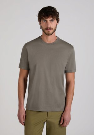 Hombre con cabello corto y rizado y barba, lleva una camiseta lisa de manga corta color verde oliva y pantalones caqui, de pie frente a un fondo gris claro.