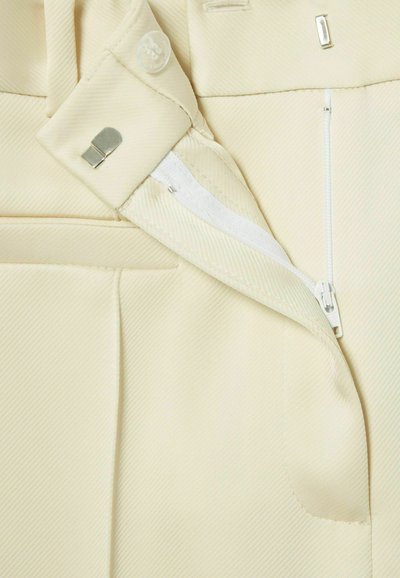 Tissu beige avec une texture côtelée, présentant une fermeture à bouton, un clip en métal et une fermeture éclair sur le côté ; comprend une poche cousue.