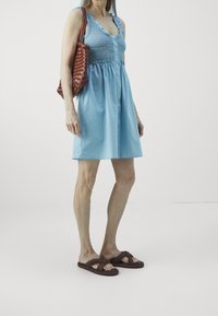 Light blue sleeveless dress with ruffled edges, cinched waist; paired with brown woven sandals and a textured brown tote bag.