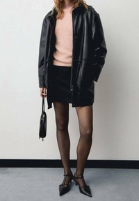 Donna che indossa una giacca di pelle nera, un maglione rosa, una mini gonna nera, collant neri trasparenti, tacchi a punta neri e tiene in mano una borsa nera.