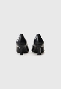 Paire de chaussures en cuir noir à talon moyen, vues de dos sur un fond clair uni.