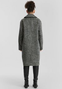 Vrouw met een krullend haar in een knot, gekleed in een lange grijs-geruite jas en zwarte kniehoge laarzen, staande met haar rug naar de camera tegen een effen achtergrond.