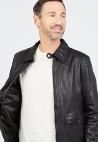 Zwarte leren jas met een kraag, ritssluiting en een voorzak. De binnenzijde is afgewerkt met een gladde stof. G gedragen over een witte trui.