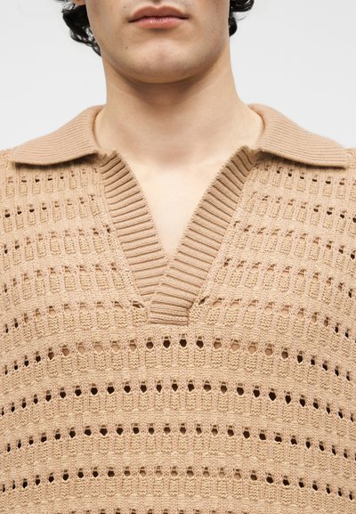 Κοντινό πλάνο ενός ατόμου που φοράει μπεζ πλεκτό πουλόβερ με ανοιχτή πλέξη και λαιμόκοψη V με ριμπ ύφανση και διπλωμένο γιακά.