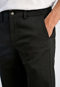 Hombre con pantalones negros y una mano en el bolsillo delantero derecho, mostrando el detalle del botón y la cintura elástica debajo de una camisa blanca.