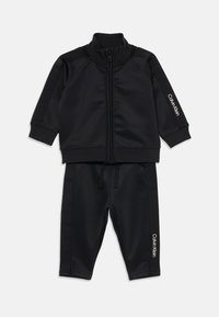 Čierny zip-up teplákový komplet vyrobený z hladkej, mäkkej tkaniny. Obsahuje rebrované manžety a lem, s bielym logom "Calvin Klein" na rukáve a nohaviciach.