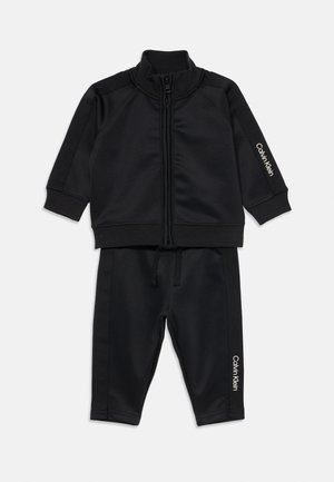 Trening negru cu fermoar, fabricat dintr-un material neted și moale. Dispune de manșete și tiv ribbate, cu branding alb "Calvin Klein" pe mânecă și pe pantalon.