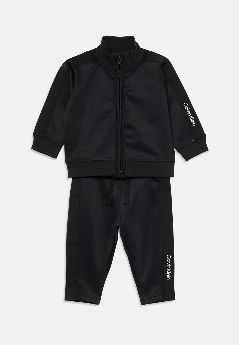 Čierny zip-up teplákový komplet vyrobený z hladkej, mäkkej tkaniny. Obsahuje rebrované manžety a lem, s bielym logom "Calvin Klein" na rukáve a nohaviciach.