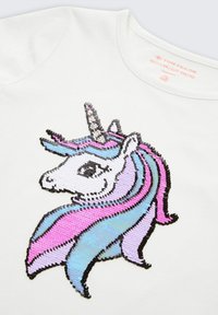 Bela majica s motivom unicornov, okrašena mavričastimi bleščicami v roza, modri in črni barvi, s srebrnim rogom in ter tečejočo grivo.