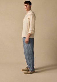 Beige, strukturierte Bluse mit zwei Vorderaschen, kombiniert mit blauen gestreiften Hosen. Das Modell trägt tanfarbene Schuhe mit gewebten Details. Der Hintergrund ist neutral.