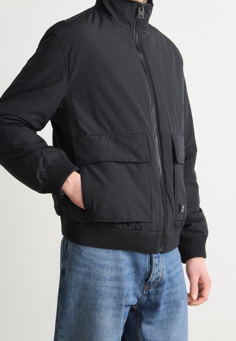 Person iført en sort jakke med lynlås, lommer med klap og ribstrikkede manchetter, kombineret med blå denimjeans, hånd i venstre lomme.