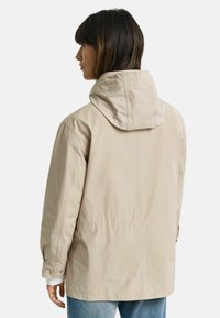 Beige hættejakke med en let oversize pasform, der har en snøret talje og knappede manchetter. Glat stof, minimalistisk design.
