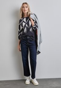 Schwarz-weißer Oberteil mit geometrischem Muster, dunkelblaue Hose und weiße Sneakers. Eine hellgraue Strickjacke ist lässig über die Schulter des Models gelegt.