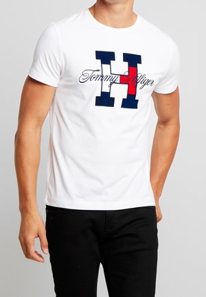 Mann, der ein weißes kurzärmliges T-Shirt mit großem navyblauem und rotem "H" sowie dem Aufdruck "Tommy Hilfiger" auf der Vorderseite trägt, kombiniert mit schwarzen Hosen.