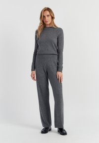 Grau gestrickter Pullover kombiniert mit grauen, weit geschnittenen Hosen, die über gerippte Bündchen und Nähte verfügen, aus einem weichen, strukturierten Stoff. Schwarze Schuhe vervollständigen den Look.