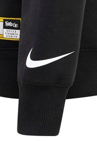 Felpa nera con polsini a costine, dotata di un logo swoosh Nike bianco e un'etichetta gialla con la scritta "Fired Up" e testo in bianco e nero.