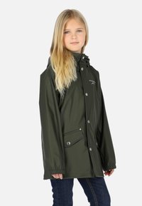 Jeune fille aux longs cheveux blonds portant un imperméable vert foncé avec des boutons-pression et des poches avant, debout devant un fond blanc uni.