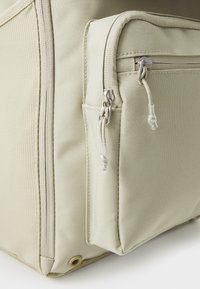 Beige fabric bag with a textured surface, featuring a zippered side pocket and silver hardware. Sturdy rectangular shape with reinforced corners.