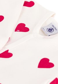 Tutina bianca in cotone con motivi a cuore rossi, dotata di chiusura a scatto sul colletto. Include un'etichetta con informazioni sulla taglia e sulla marca.