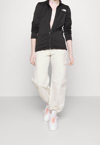 Czarna polarowa kurtka na zamek, jasnoróżowy t-shirt z grafiką pod spodem, jasne spodnie cargo i białe trampki z pomarańczowymi akcentami.