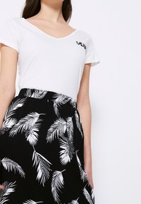 Nainen, joka pukeutuu valkoiseen V-kaula-aukkoiseen T-paitaan, jossa on pientä mustaa tekstiä rinnassa, ja mustaan hameeseen, jossa on valkoinen trooppinen lehtikuvio.