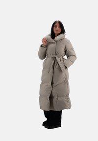 Lang, puffy frakke i lys beige med høj krave, bælte i taljen og store quiltede sektioner. Bæres over sorte bukser og sko.