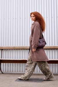 Hnedý dlhý kabát cez svetlý top, vzorované šedé široké nohavice a fialová crossbody taška. Kučeravé červené vlasy, kráčajúca po chodníku.