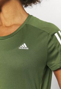 Camiseta deportiva verde oliva con cuello redondo, tela texturizada y logo blanco de Adidas. Presenta paneles laterales de malla y tres franjas blancas en las mangas.