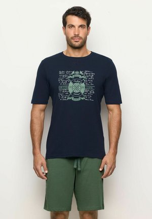 Hombre vistiendo una camiseta de manga corta azul marino con un emblema de navegación y pantalones cortos verdes, de pie contra un fondo claro y liso.