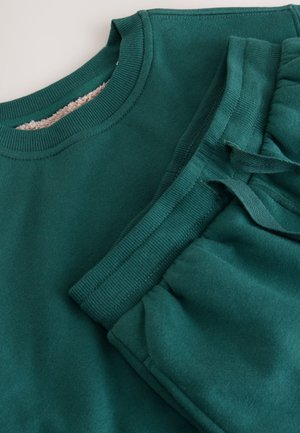 Grüner Pullover und passende Sweatpants mit gerippten Bündchen und weicher Textur, seitlichen Taschen und einem Kordelzug an der Taille der Hose.