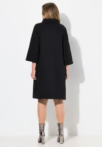 Vestido negro de gran tamaño con un cuello alto, mangas de tres cuartos y textura suave, combinado con botas metálicas hasta el tobillo y un acabado liso.
