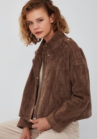 Bruine suede jas met een ritssluiting, drukknoopaccenten, zakken aan de zijkant en een relaxte pasvorm. Voorzien van een kraag en een gestructureerd oppervlak.