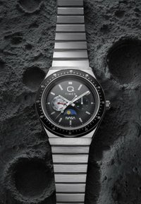 Zegarek Timex ze stali nierdzewnej z logo NASA, tarczkami dni i sekund, wyświetlany na teksturowanym tle powierzchni Księżyca.
