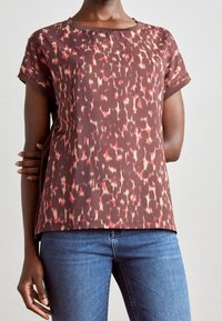 Mujer vistiendo una blusa marrón de mangas cortas con manchas abstractas rojas y beige y jeans de denim azul, contra un fondo liso.