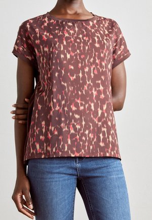 Donna che indossa una maglietta a maniche corte marrone con macchie astratte rosse e beige e jeans di denim blu su uno sfondo semplice.