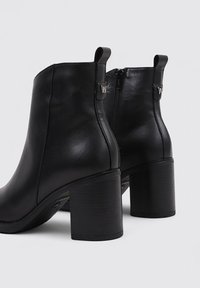 Botas de tobillo de cuero negro con punta redonda, cremallera lateral y tacón cuadrado. Presenta una lengüeta en la parte posterior con un acento de logotipo.