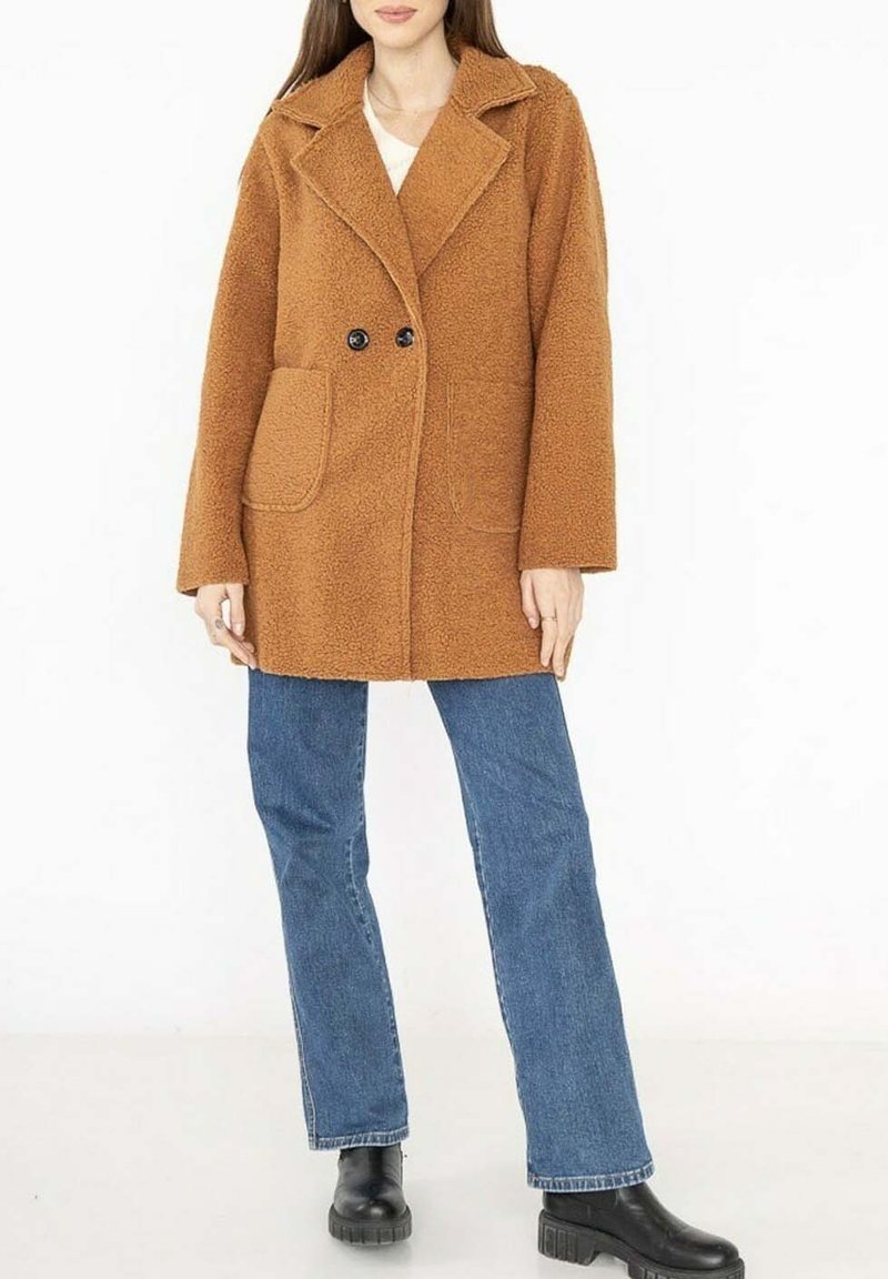 Un manteau en peluche brun avec un col à revers, deux boutons noirs et des poches latérales. Associé à un jean bleu et des bottines noires.