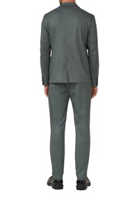 Homme portant un costume gris ajusté avec des manches longues et un pantalon assorti, se tenant droit avec des chaussures de ville noires visibles.