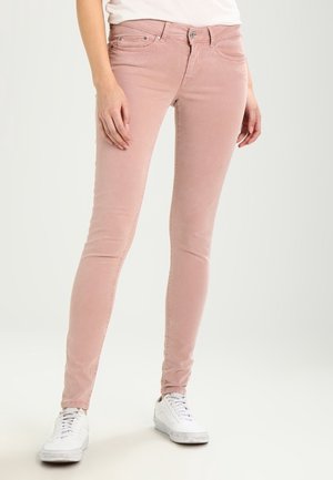 Frau trägt hellrosa Skinny-Jeans und weiße Sneaker, steht mit entspannt an den Seiten hängenden Händen vor einfachem Hintergrund.